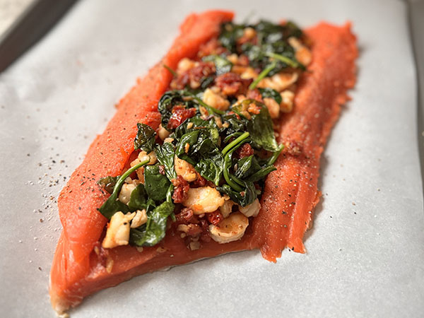 Salmon filet with shrimp, sun-dried tomatoes and spinach filling on top, on a parchment paper covered baking sheet.