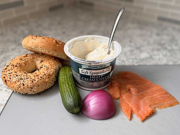 Ингредиенты для бейгл-сэндвича на разделочной доске: бейглы, сливочный сыр, огурец, красный лук и копченый лосось.