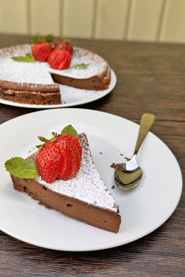 A piece of chocolate cake topped with powdered sugar, strawberry and mint, with the rect of the cake in the background.