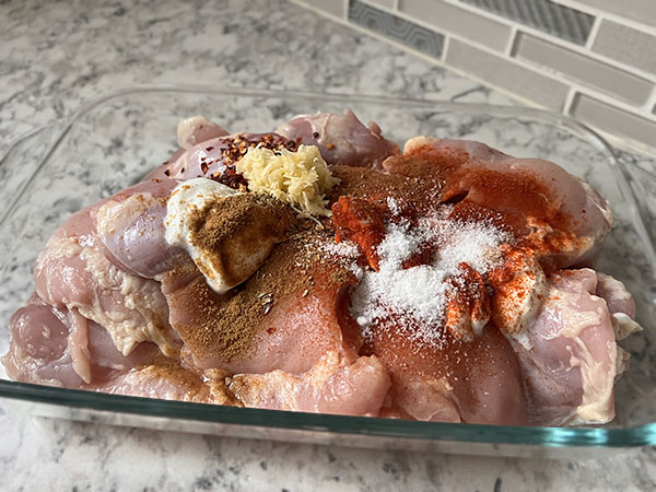 Chicken thighs in a glass dish with shawarma marinade ingredients on top.