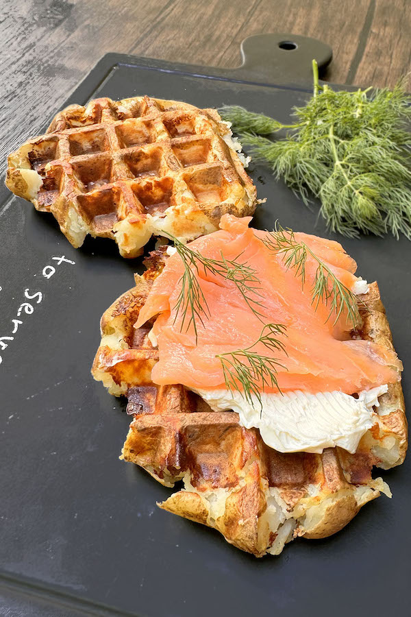 Smashed potato waffle topped with cream cheese and smoked salmon with another potato waffle and fresh dill on the background.