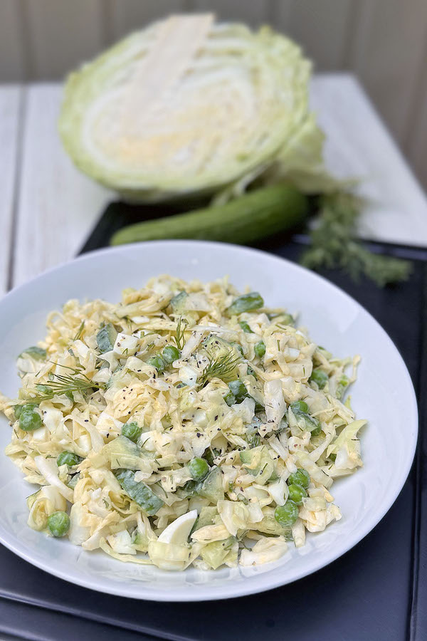 Serving of fresh cabbage salad with peas and creamy Dijon dressing with half head of cabbage, cucumber and dill in the background.