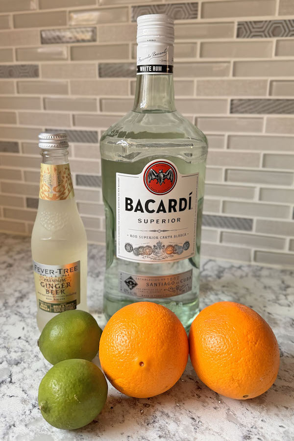 Ingredients for orange Moscow mule: Bacardi rum, ginger beer, limes and oranges on a countertop.