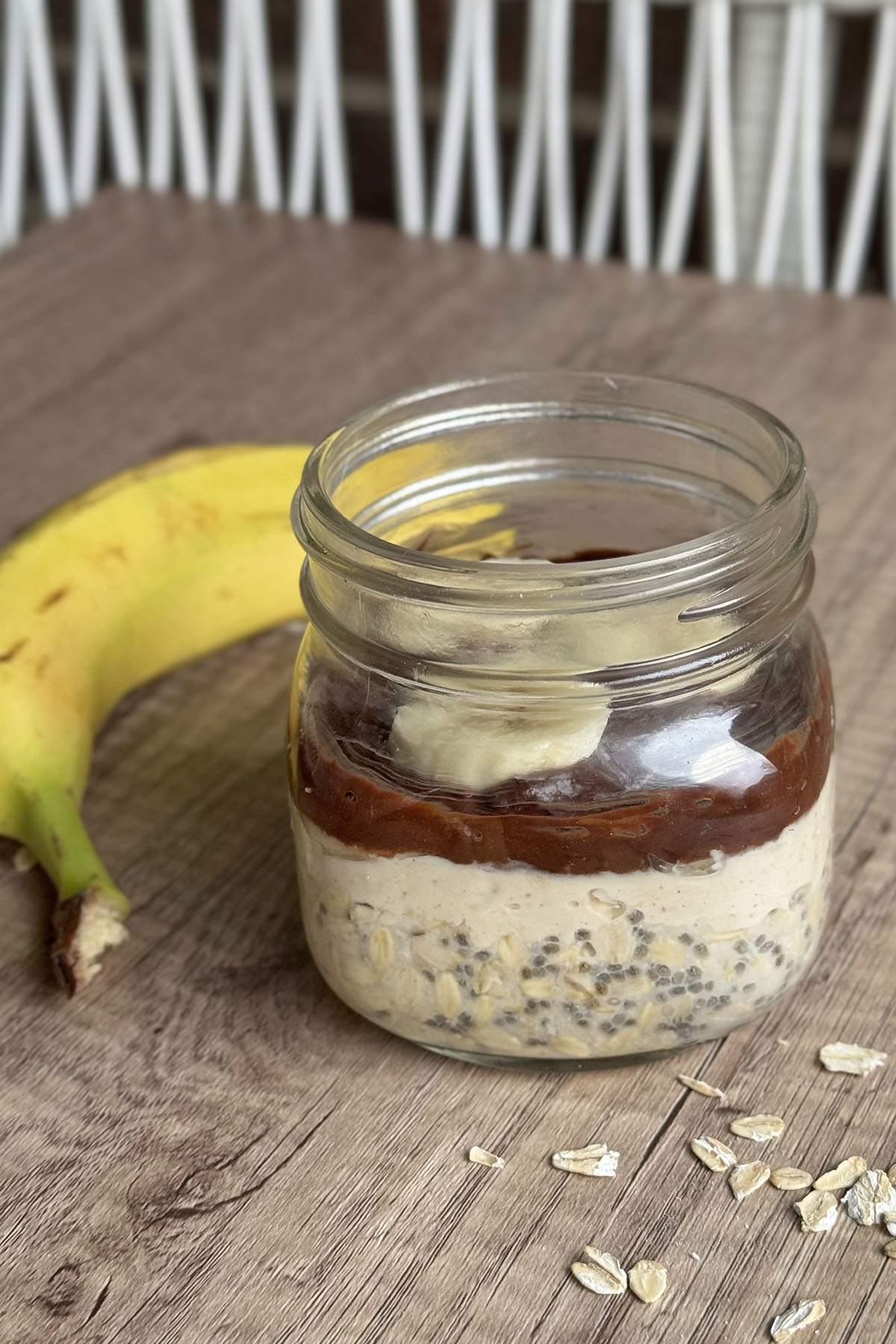 Protein-packed peanut butter chocolate oats breakfast jar, topped with banana slices with a banana on the background.