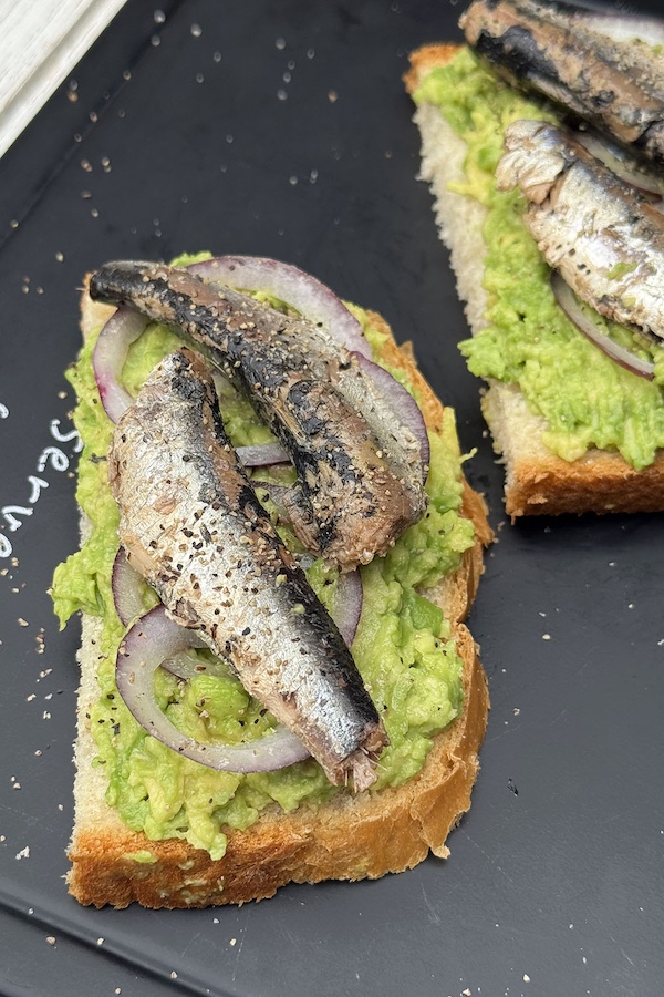 Two avocado and canned sardines sandwiches on a serving board sprinkled with salt and black pepper.